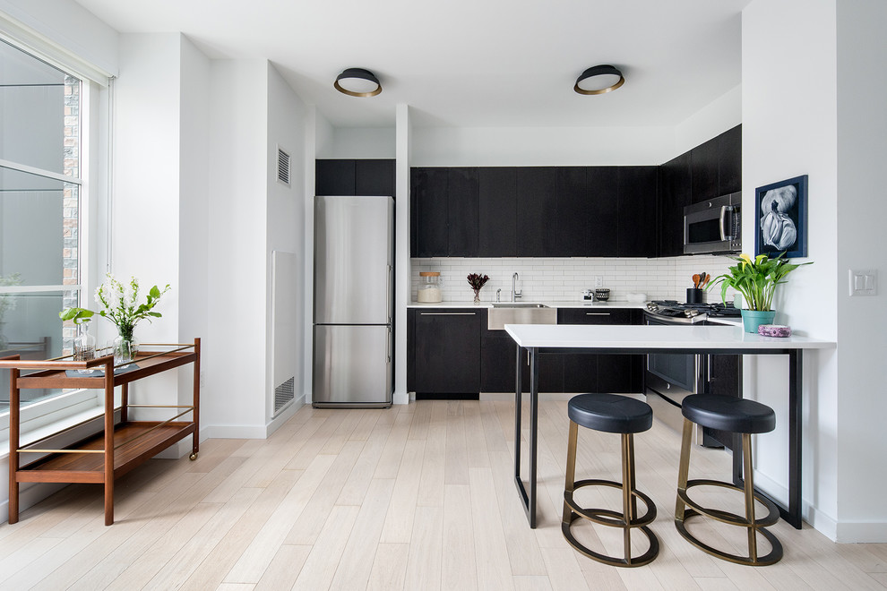 Design ideas for a contemporary l-shaped kitchen in New York with a farmhouse sink, flat-panel cabinets, black cabinets, white splashback, subway tile splashback, stainless steel appliances, light hardwood floors, a peninsula and beige floor.