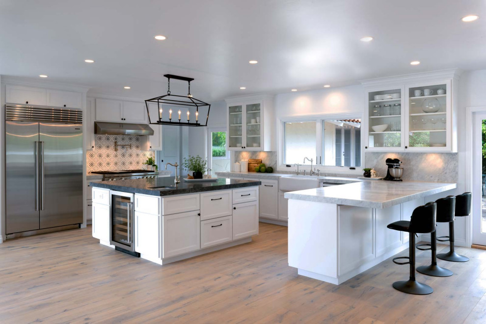 Large country u-shaped eat-in kitchen in Santa Barbara with a farmhouse sink, white cabinets, marble benchtops, grey splashback, ceramic splashback, stainless steel appliances, light hardwood floors, beige floor and grey benchtop.