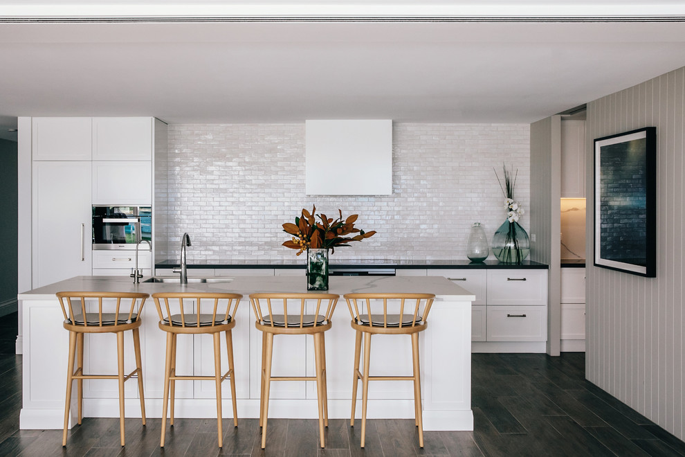 This is an example of a beach style galley kitchen in Sydney with an undermount sink, shaker cabinets, white cabinets, white splashback, panelled appliances, dark hardwood floors, with island, brown floor and black benchtop.