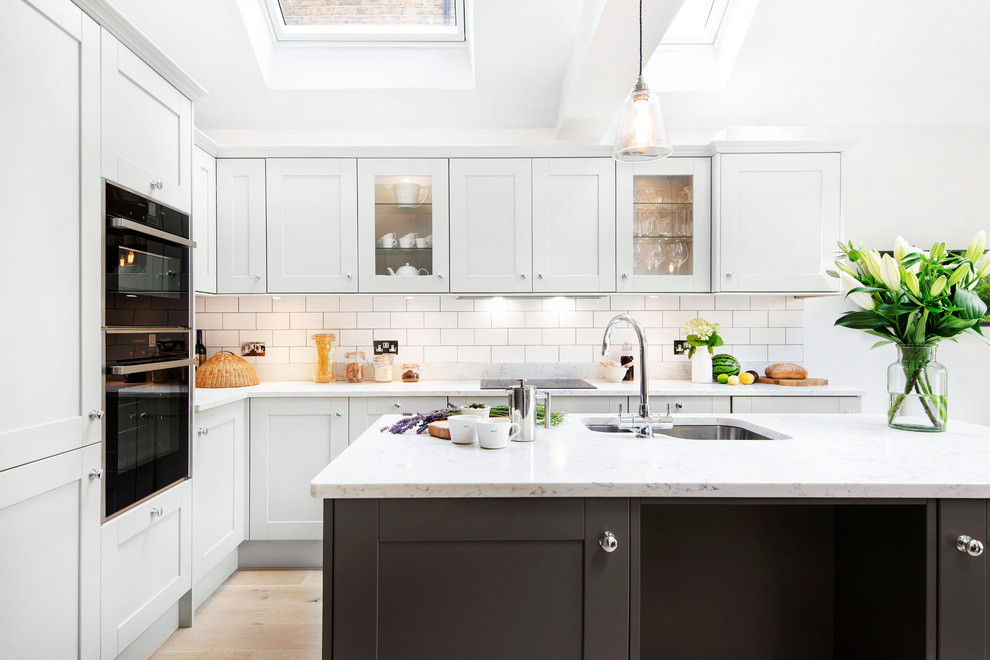 Design ideas for a mid-sized transitional l-shaped kitchen in London with shaker cabinets, grey cabinets, quartz benchtops, white splashback, black appliances, light hardwood floors, with island, white benchtop, an undermount sink and subway tile splashback.