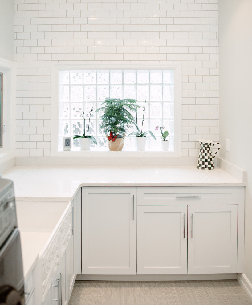 This is an example of a transitional laundry room in Detroit with a farmhouse sink, shaker cabinets, blue cabinets, quartz benchtops, white splashback, subway tile splashback, grey walls, porcelain floors, a side-by-side washer and dryer and white benchtop.