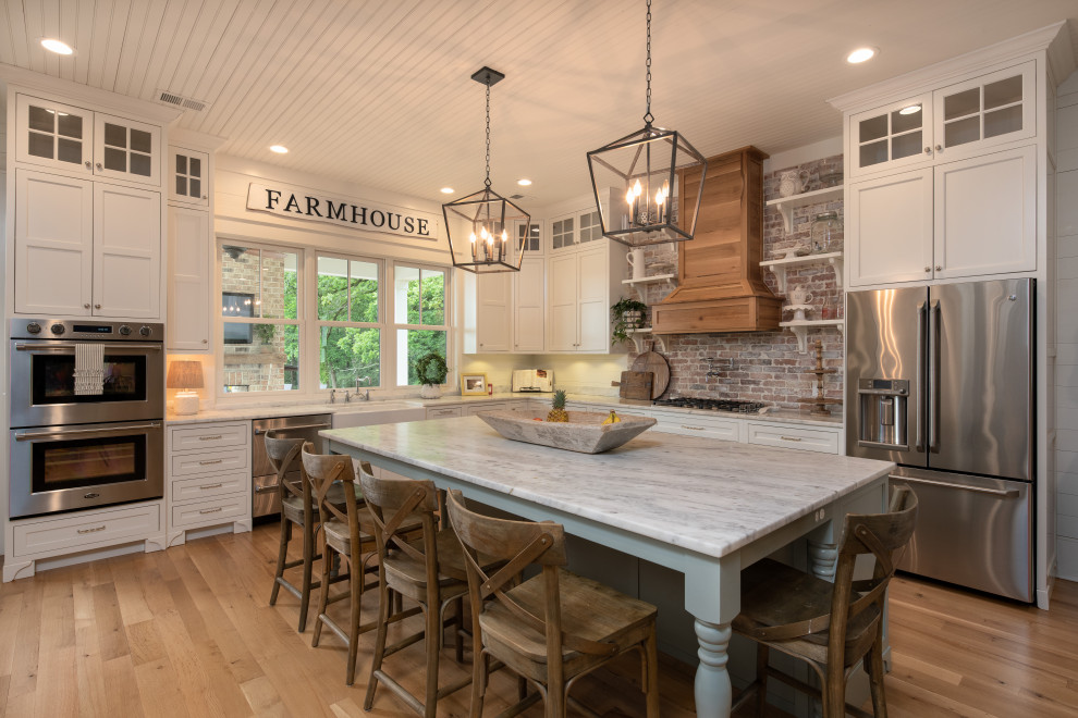 This is an example of a large country l-shaped eat-in kitchen in Other with a farmhouse sink, flat-panel cabinets, white cabinets, granite benchtops, brown splashback, brick splashback, stainless steel appliances, light hardwood floors, with island, beige floor and white benchtop.