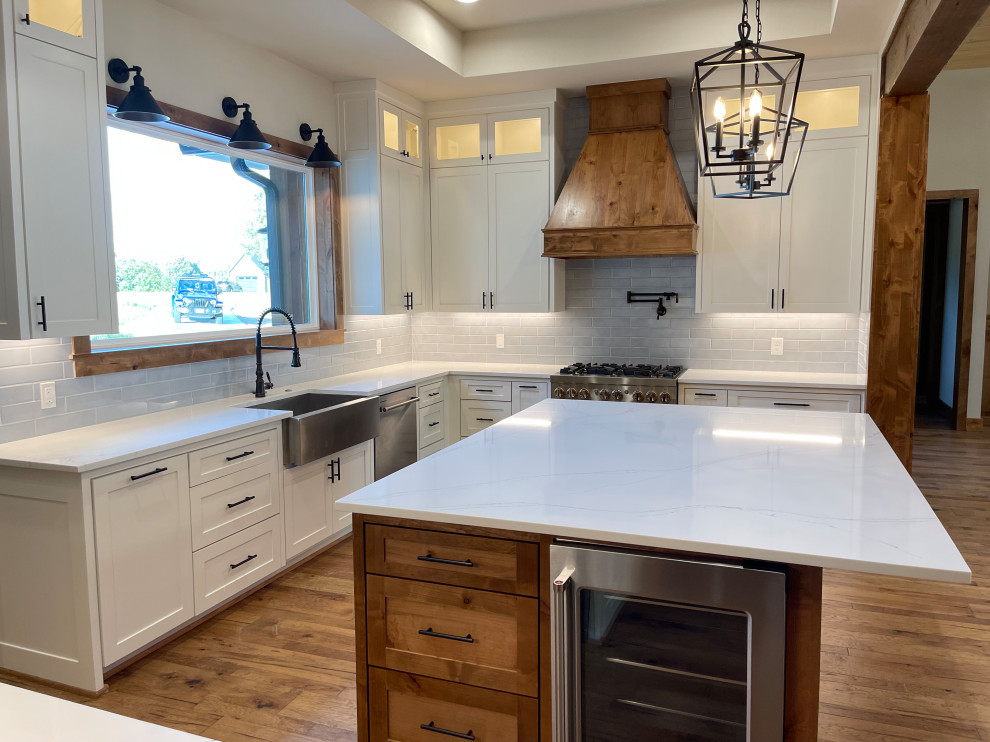 This is an example of a large arts and crafts l-shaped kitchen in Dallas with a farmhouse sink, shaker cabinets, white cabinets, quartz benchtops, blue splashback, cement tile splashback, stainless steel appliances, light hardwood floors, with island, brown floor, white benchtop and recessed.