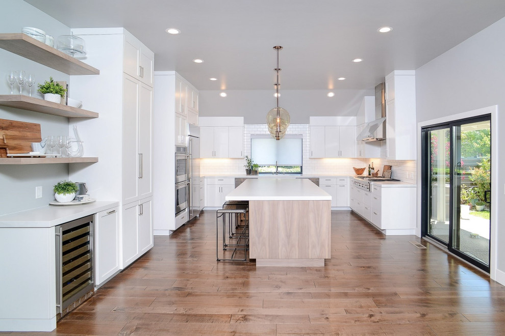 Photo of a transitional u-shaped open plan kitchen in Sacramento with shaker cabinets, white cabinets, white splashback, subway tile splashback, stainless steel appliances, medium hardwood floors, with island, brown floor and white benchtop.