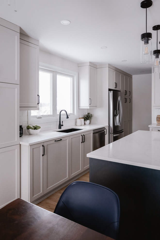 Mid-sized transitional l-shaped light wood floor and brown floor eat-in kitchen photo with an undermount sink, shaker cabinets, beige cabinets, quartz countertops, white backsplash, subway tile backsplash, stainless steel appliances, an island and white countertops