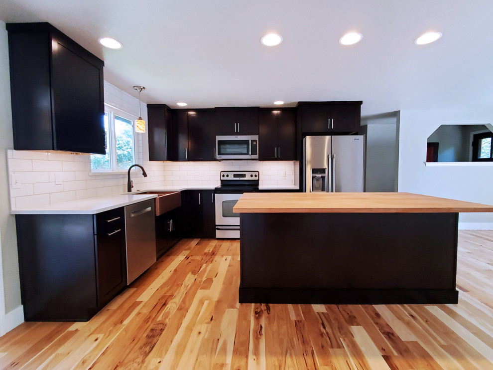 Eat-in kitchen - mid-sized transitional l-shaped medium tone wood floor eat-in kitchen idea in Boise with a farmhouse sink, shaker cabinets, black cabinets, quartz countertops, white backsplash, porcelain backsplash, stainless steel appliances, an island and white countertops