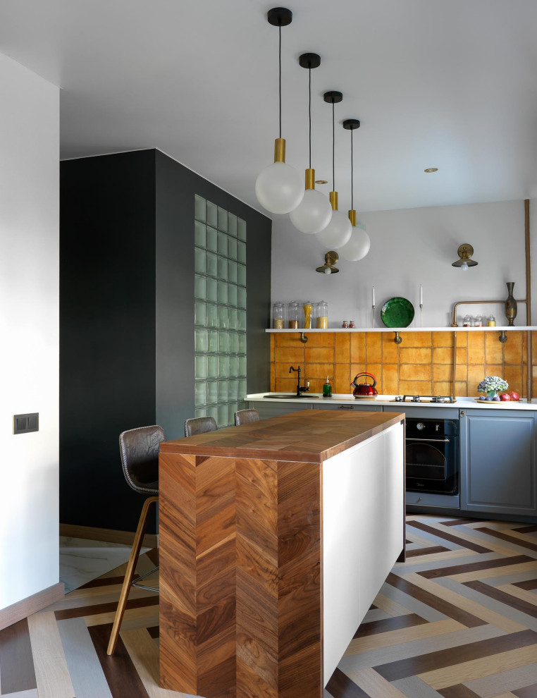 Inspiration for a small eclectic single-wall open plan kitchen in Moscow with a drop-in sink, recessed-panel cabinets, grey cabinets, solid surface benchtops, orange splashback, ceramic splashback, black appliances, medium hardwood floors, with island, multi-coloured floor and white benchtop.