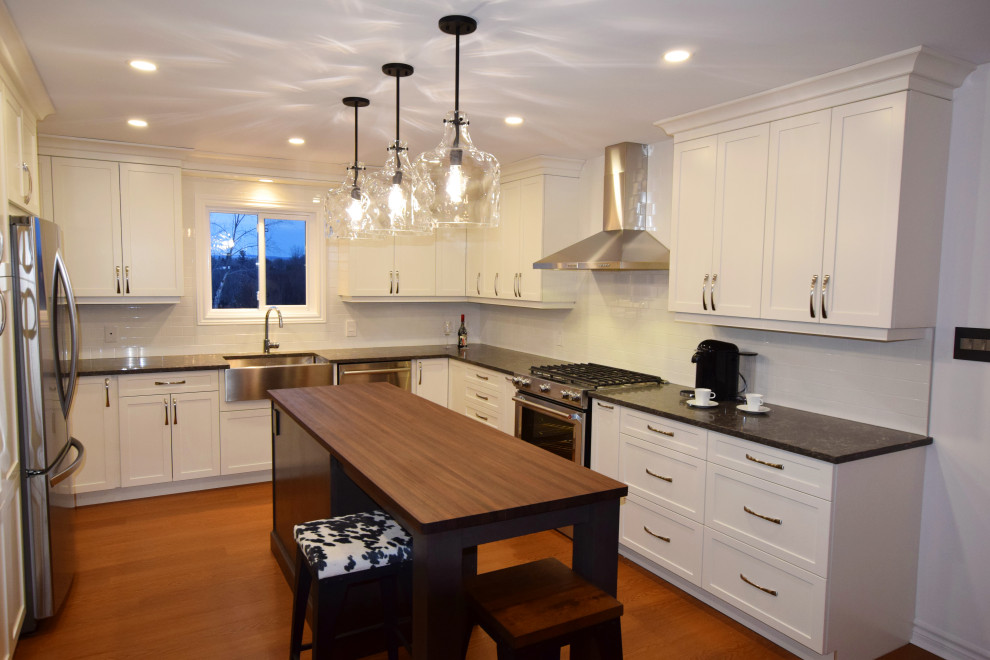 Example of a small transitional u-shaped medium tone wood floor and brown floor open concept kitchen design in Ottawa with a farmhouse sink, shaker cabinets, white cabinets, quartz countertops, white backsplash, subway tile backsplash, stainless steel appliances, an island and gray countertops