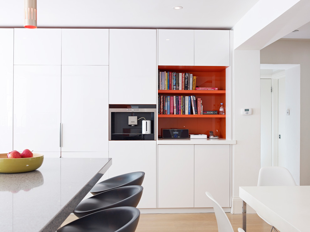 This is an example of a mid-sized contemporary eat-in kitchen in London with flat-panel cabinets, white cabinets, solid surface benchtops, orange splashback, glass sheet splashback, black appliances and with island.