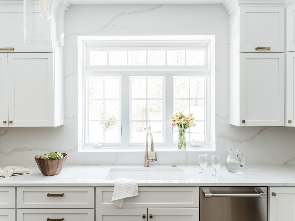 This is an example of a transitional u-shaped kitchen in Providence with an undermount sink, recessed-panel cabinets, white cabinets, quartz benchtops, white splashback, engineered quartz splashback, stainless steel appliances, light hardwood floors, with island and white benchtop.