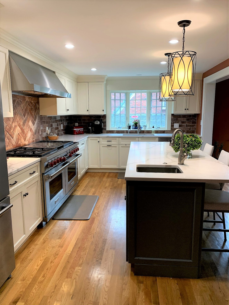 Large classic l-shaped kitchen pantry in Boston with a submerged sink, shaker cabinets, white cabinets, engineered stone countertops, brown splashback, brick splashback, stainless steel appliances, light hardwood flooring, an island, brown floors and white worktops.