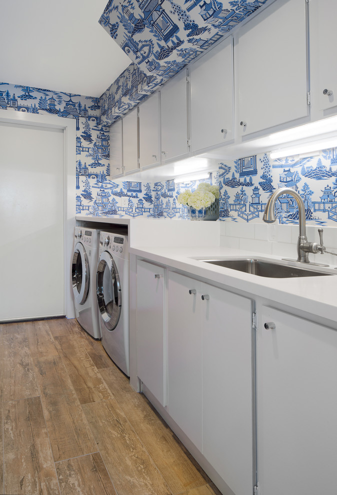 This is an example of a transitional single-wall laundry room in San Diego with an undermount sink, flat-panel cabinets, white cabinets, blue walls, medium hardwood floors, a side-by-side washer and dryer and white benchtop.