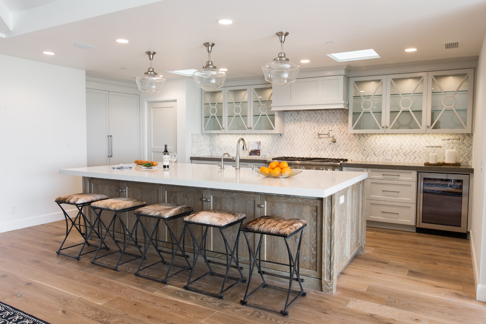 Mid-sized beach style galley eat-in kitchen in Orange County with a farmhouse sink, glass-front cabinets, soapstone benchtops, white splashback, mosaic tile splashback, stainless steel appliances, with island, light hardwood floors, grey cabinets and beige floor.