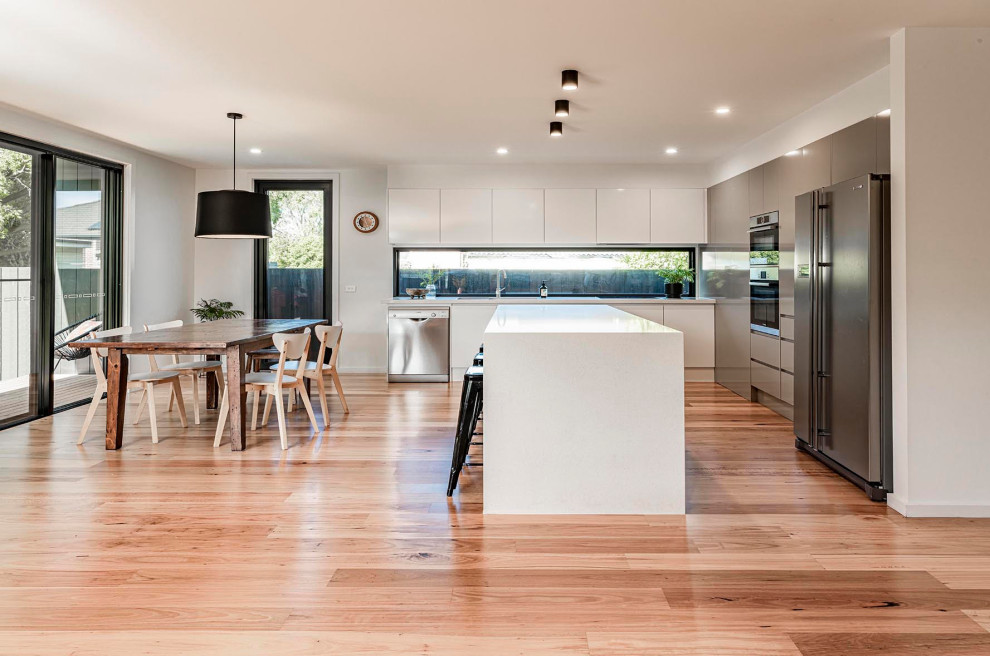 Expansive contemporary l-shaped kitchen in Melbourne with flat-panel cabinets, white cabinets, mirror splashback, stainless steel appliances, medium hardwood floors, with island, beige floor and white benchtop.