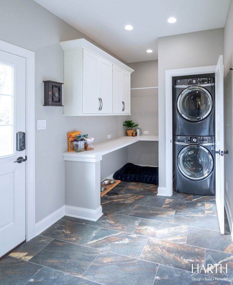 Photo of a mid-sized transitional l-shaped utility room in Philadelphia with shaker cabinets, white cabinets, wood benchtops, grey walls, porcelain floors, a stacked washer and dryer, black floor and white benchtop.