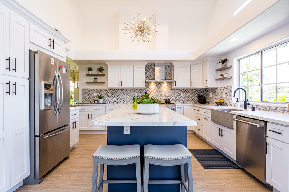 Large beach style l-shaped eat-in kitchen in Los Angeles with a farmhouse sink, shaker cabinets, blue cabinets, solid surface benchtops, grey splashback, ceramic splashback, stainless steel appliances, medium hardwood floors, with island, brown floor, white benchtop and vaulted.