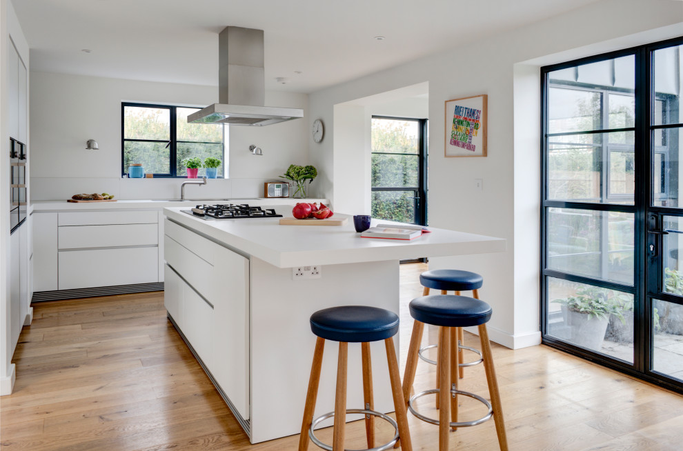 Design ideas for a modern l-shaped eat-in kitchen in Gloucestershire with an integrated sink, flat-panel cabinets, white cabinets, laminate benchtops, white splashback, glass sheet splashback, black appliances, medium hardwood floors, with island and white benchtop.