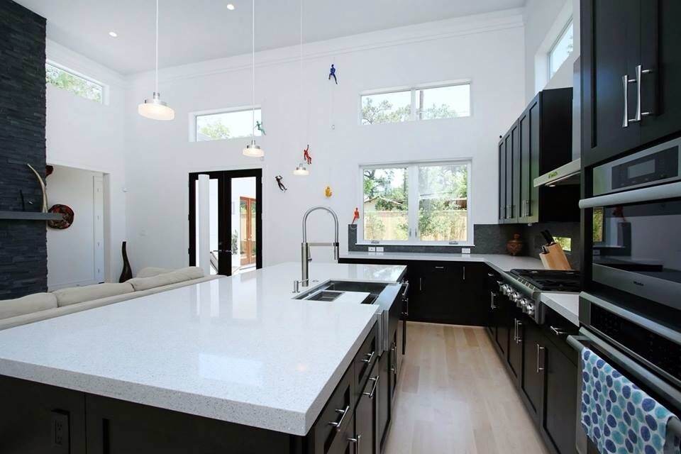 Modern kitchen in Houston with with island, black cabinets, light hardwood floors, a farmhouse sink, quartz benchtops, black splashback, limestone splashback and stainless steel appliances.