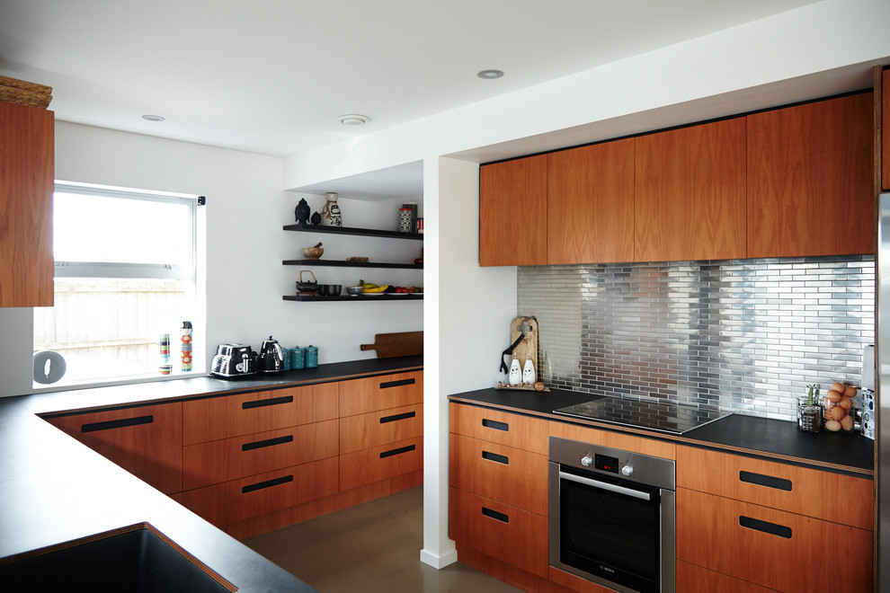 Inspiration for an expansive contemporary galley kitchen pantry in Christchurch with an undermount sink, flat-panel cabinets, medium wood cabinets, solid surface benchtops, metallic splashback, metal splashback, stainless steel appliances, concrete floors, a peninsula, beige floor and black benchtop.