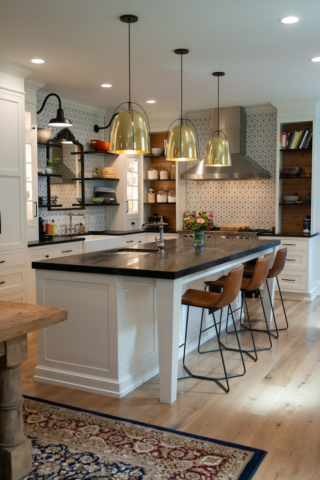 Photo of a transitional l-shaped kitchen in Philadelphia with soapstone benchtops, black benchtop, a farmhouse sink, recessed-panel cabinets, white cabinets, white splashback, mosaic tile splashback, light hardwood floors, with island and beige floor.