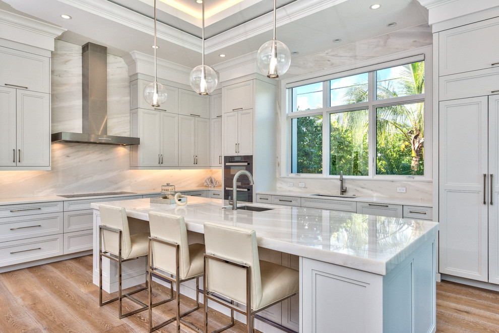 Design ideas for a beach style l-shaped kitchen in Miami with an undermount sink, recessed-panel cabinets, white cabinets, white splashback, stainless steel appliances, medium hardwood floors, with island, brown floor and white benchtop.
