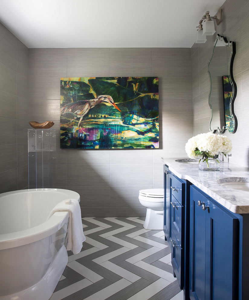 This is an example of a mid-sized transitional master bathroom in Charleston with shaker cabinets, blue cabinets, a freestanding tub, a one-piece toilet, grey walls, an undermount sink, marble benchtops, grey floor, gray tile, porcelain tile, porcelain floors and grey benchtops.