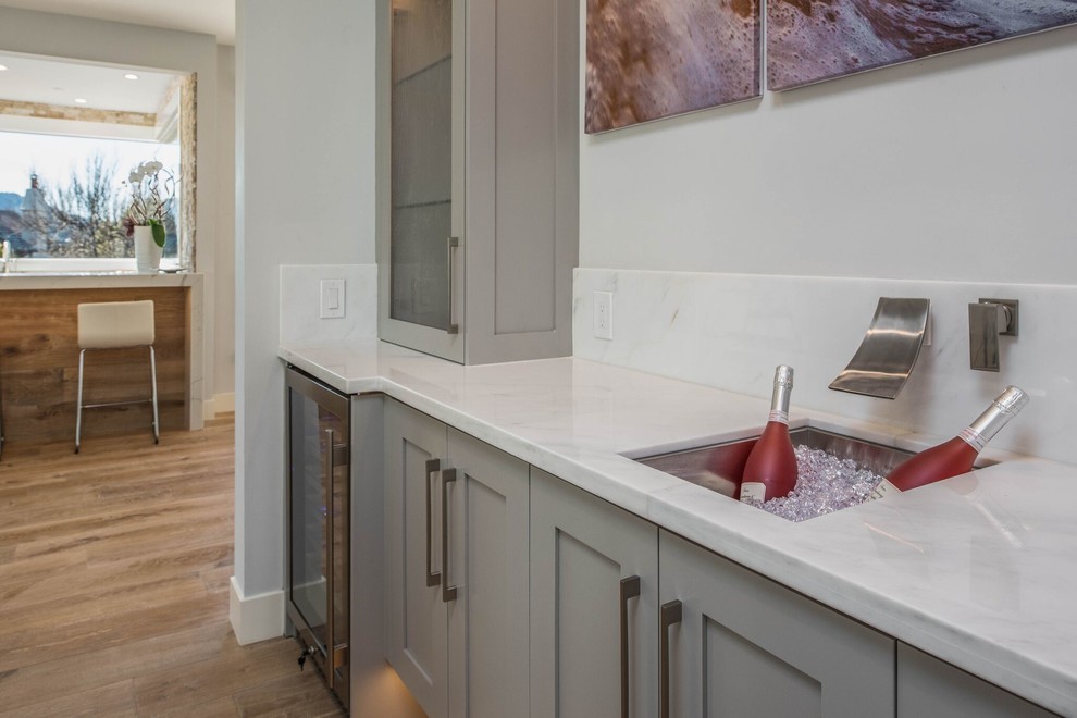 Photo of a mid-sized transitional single-wall wet bar in Orange County with an undermount sink, glass-front cabinets, grey cabinets, quartzite benchtops, light hardwood floors and white benchtop.