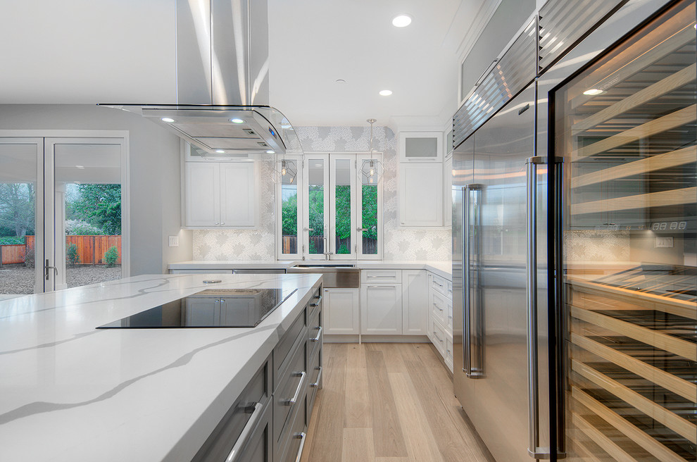 Photo of a large transitional l-shaped kitchen in San Francisco with a farmhouse sink, shaker cabinets, white cabinets, white splashback, stainless steel appliances, light hardwood floors, with island and brown floor.