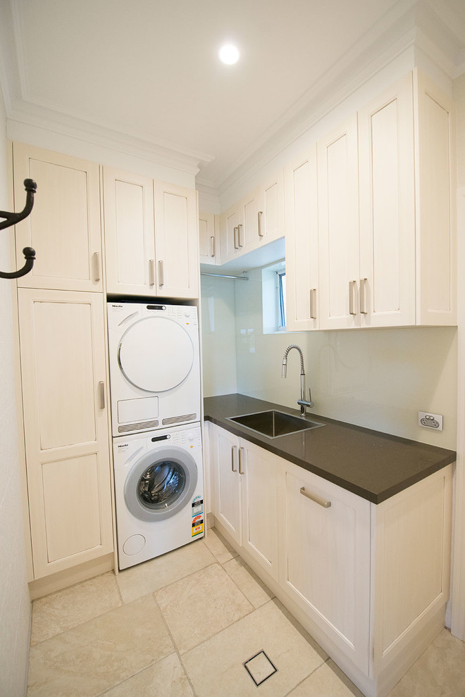 Photo of a large traditional l-shaped laundry room in Sydney with beige cabinets, white walls, a stacked washer and dryer, shaker cabinets, quartz benchtops, a farmhouse sink, beige splashback, glass sheet splashback and travertine floors.