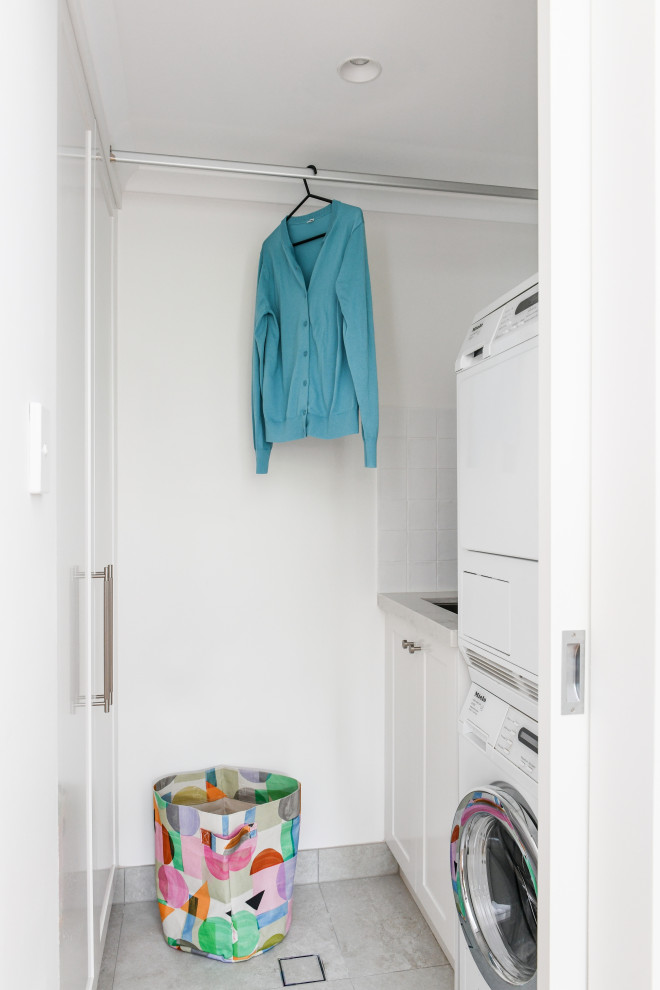 Inspiration for a small traditional single-wall dedicated laundry room in Sydney with an undermount sink, white cabinets, quartz benchtops, white splashback, ceramic splashback, white walls, ceramic floors, a stacked washer and dryer, grey floor and white benchtop.