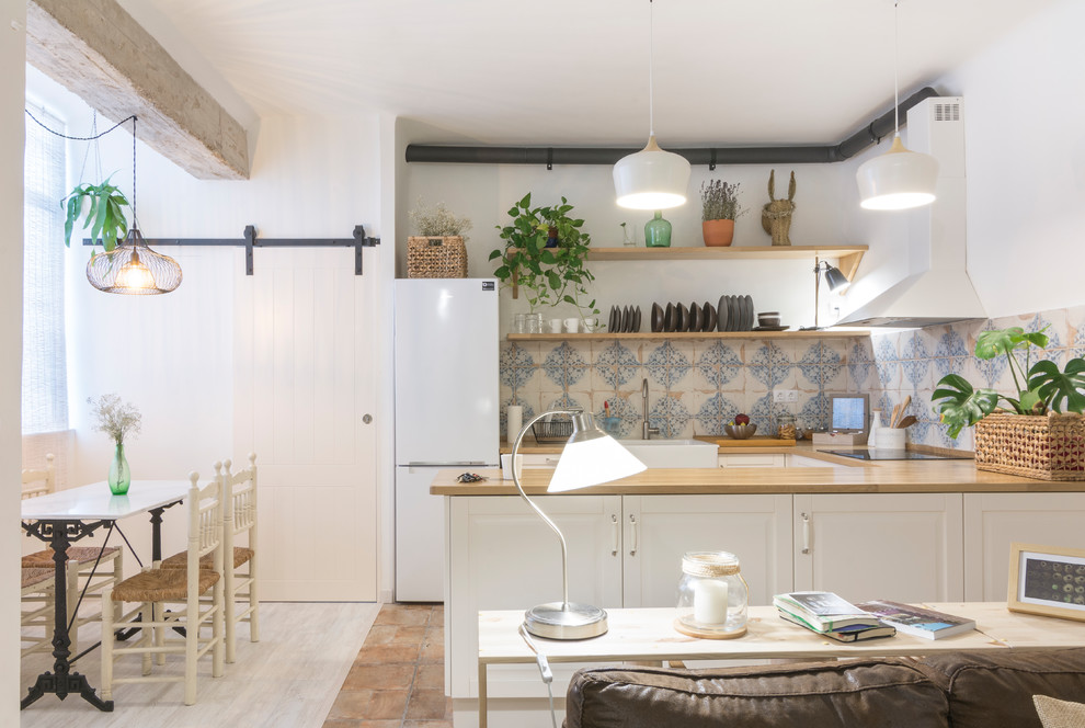 This is an example of a mid-sized country u-shaped open plan kitchen in Other with a farmhouse sink, raised-panel cabinets, white cabinets, wood benchtops, blue splashback, white appliances, terra-cotta floors, a peninsula and brown floor.