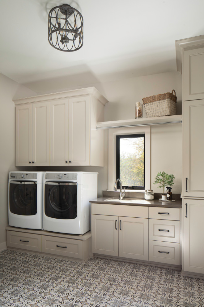 Klassisk inredning av en stor grå grått tvättstuga, med en allbänk, släta luckor, grå skåp, grå väggar, klinkergolv i keramik, en tvättmaskin och torktumlare bredvid varandra och flerfärgat golv