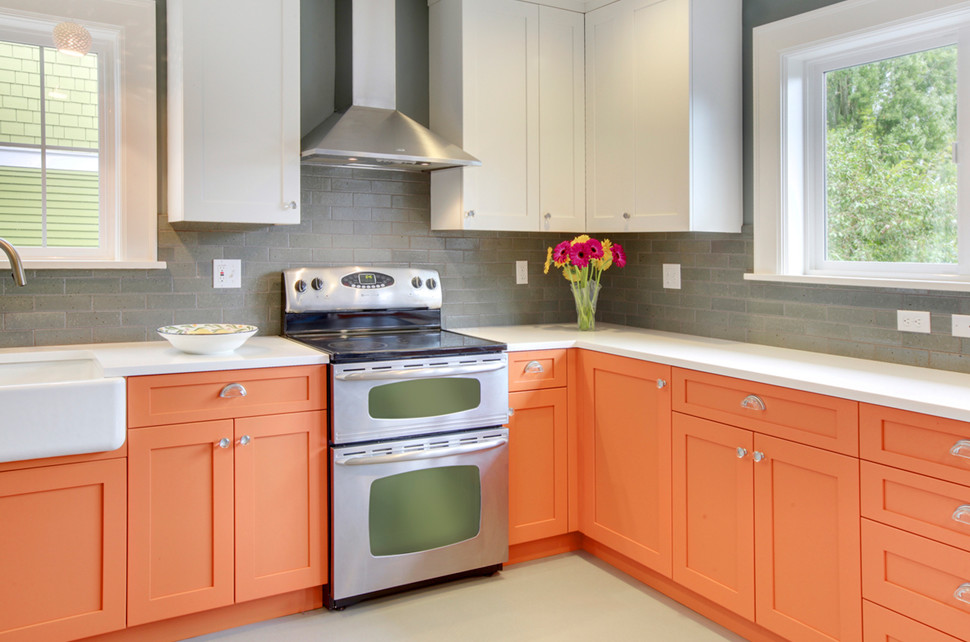 This is an example of a mid-sized modern l-shaped eat-in kitchen in Seattle with a farmhouse sink, shaker cabinets, orange cabinets, solid surface benchtops, grey splashback, subway tile splashback, stainless steel appliances, grey floor and white benchtop.