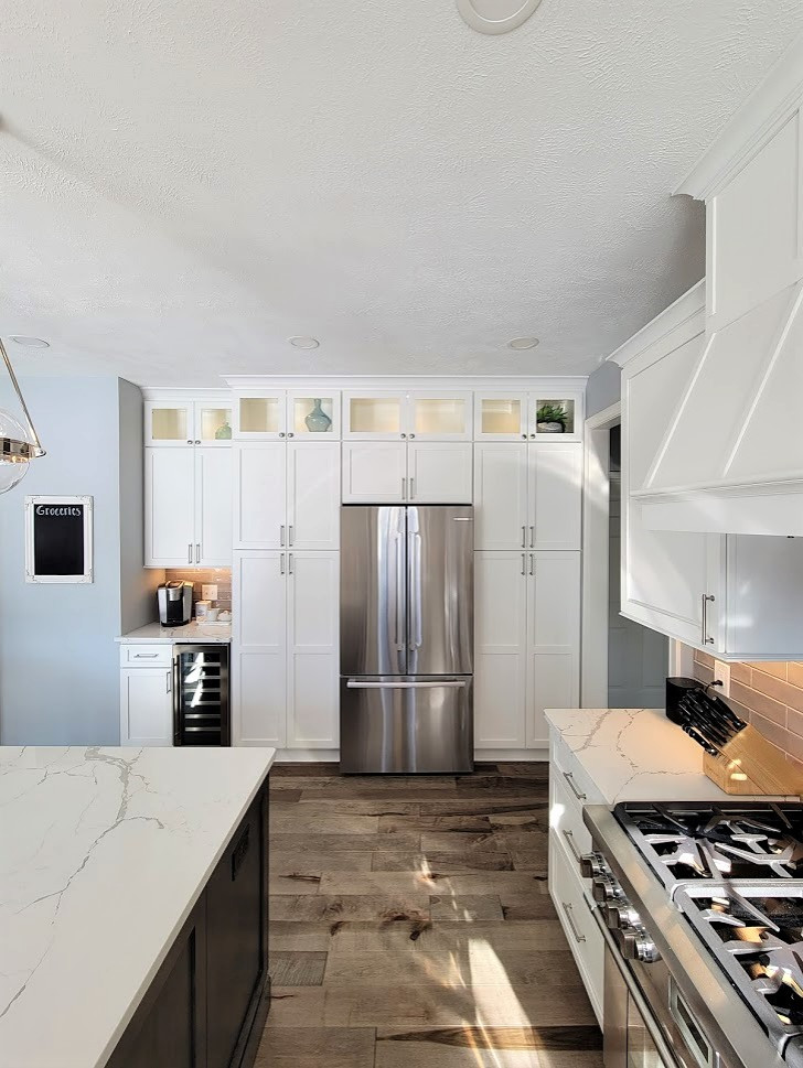 This is an example of a large transitional l-shaped eat-in kitchen in Cleveland with shaker cabinets, white cabinets, quartz benchtops, grey splashback, subway tile splashback, stainless steel appliances, light hardwood floors, with island and white benchtop.