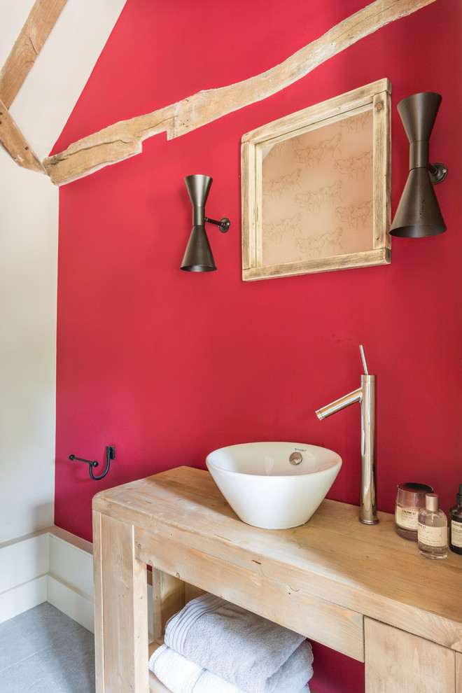 Country bathroom with light wood cabinets, red walls, a vessel sink and grey floor.