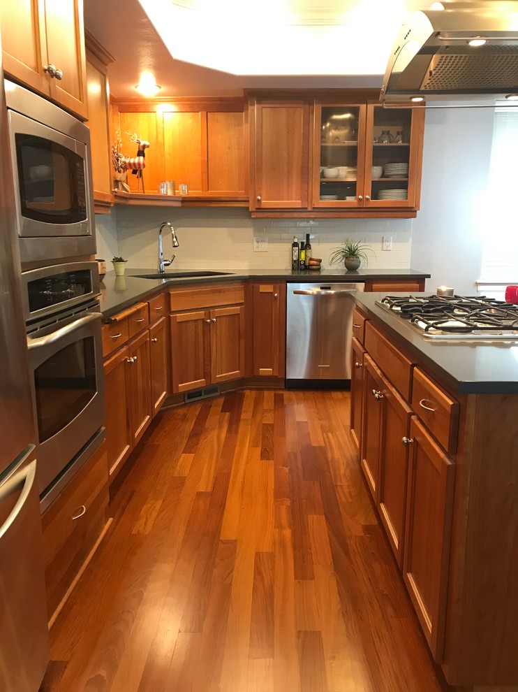 This is an example of a small arts and crafts u-shaped eat-in kitchen in Portland with an undermount sink, shaker cabinets, medium wood cabinets, quartz benchtops, white splashback, ceramic splashback, stainless steel appliances, medium hardwood floors, with island, brown floor and grey benchtop.