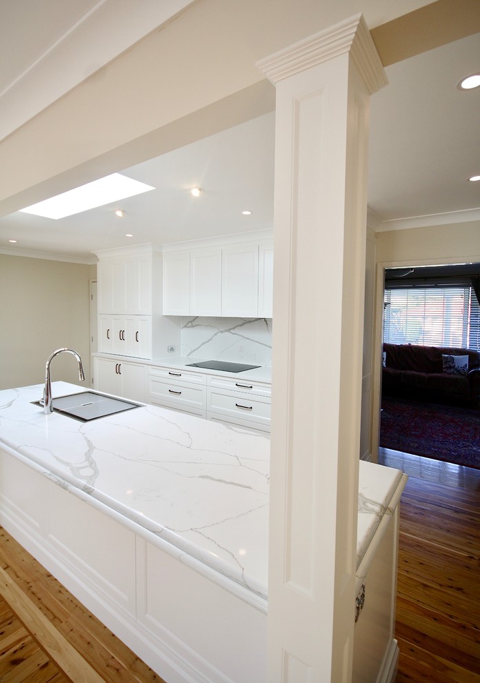 This is an example of a large transitional u-shaped open plan kitchen in Sydney with a drop-in sink, recessed-panel cabinets, white cabinets, quartz benchtops, white splashback, stone slab splashback, stainless steel appliances, medium hardwood floors, with island, brown floor and white benchtop.