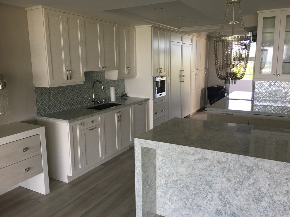 Large transitional l-shaped light wood floor and beige floor kitchen photo in Miami with an undermount sink, shaker cabinets, white cabinets, quartzite countertops, gray backsplash, glass tile backsplash, stainless steel appliances and an island