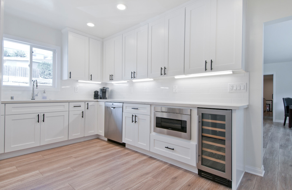 Photo of a mid-sized transitional l-shaped separate kitchen in Los Angeles with an undermount sink, shaker cabinets, white cabinets, white splashback, porcelain splashback, stainless steel appliances, light hardwood floors, no island, brown floor and white benchtop.