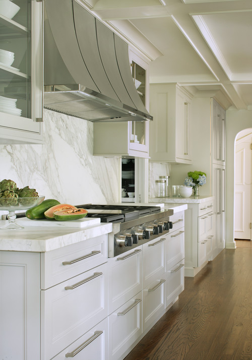 Elegant galley kitchen with white shaker RTA cabinets, long stainless bar pulls, a veined marble slab backsplash and counters, pro-style range under a curved stainless hood, and warm wood floors.