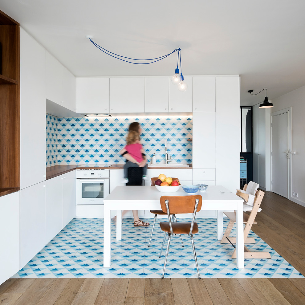 Photo of a mid-sized scandinavian l-shaped eat-in kitchen in Paris with a drop-in sink, flat-panel cabinets, white cabinets, wood benchtops, multi-coloured splashback and white appliances.