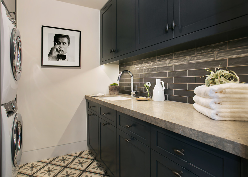 Inspiration for a transitional galley dedicated laundry room in Orange County with an undermount sink, shaker cabinets, black cabinets, white walls, a stacked washer and dryer, multi-coloured floor and grey benchtop.