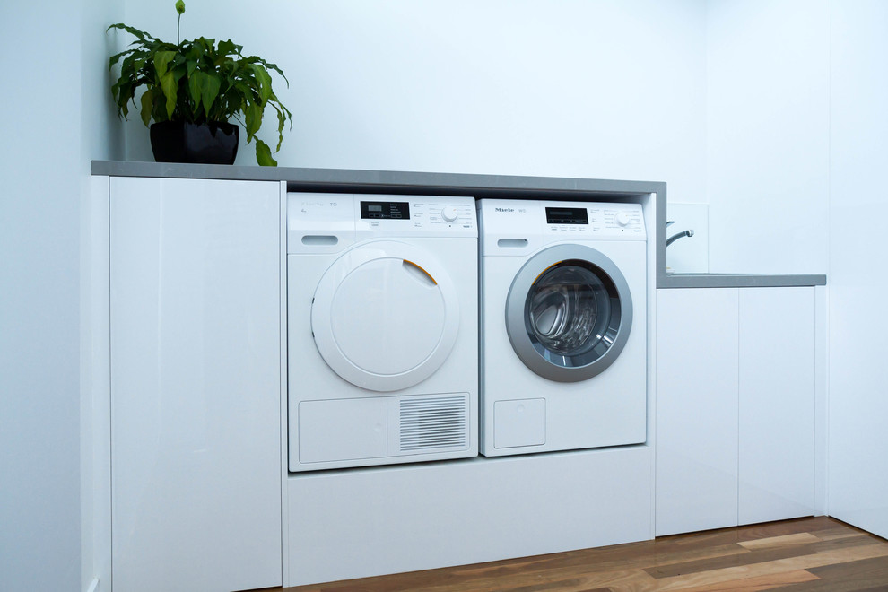 Photo of a mid-sized modern dedicated laundry room in Melbourne with flat-panel cabinets, white cabinets, quartz benchtops, white walls, medium hardwood floors and a side-by-side washer and dryer.