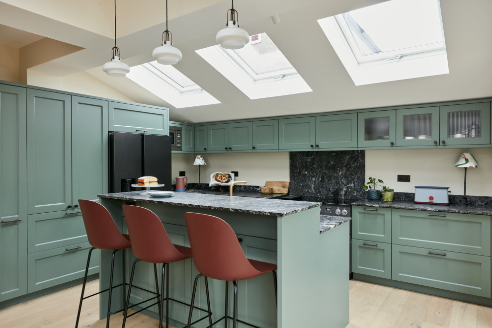 This is an example of a mid-sized traditional l-shaped open plan kitchen in London with a farmhouse sink, recessed-panel cabinets, green cabinets, granite benchtops, black splashback, granite splashback, light hardwood floors, with island, black benchtop and vaulted.