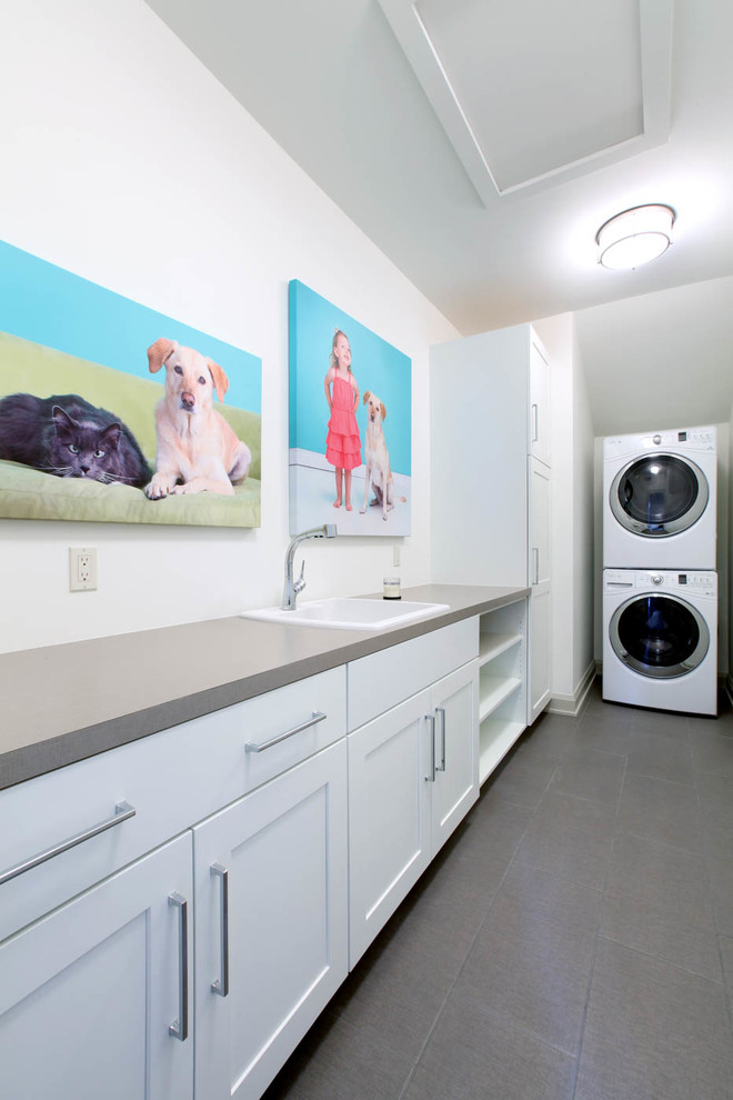 Photo of a small transitional l-shaped dedicated laundry room in Grand Rapids with a drop-in sink, shaker cabinets, white cabinets, laminate benchtops, grey walls, ceramic floors, a stacked washer and dryer, grey floor and grey benchtop.