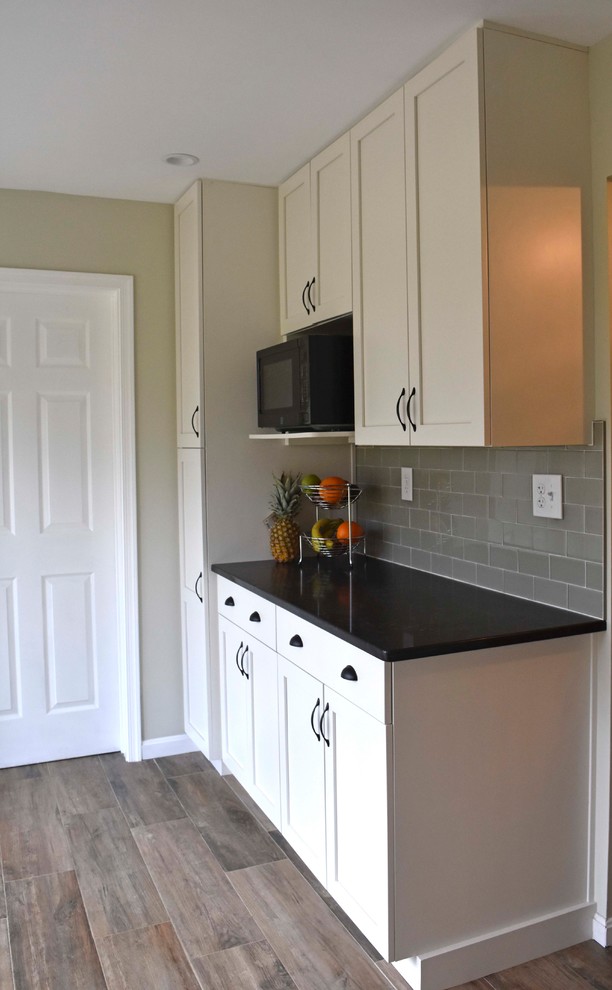 Example of a mid-sized transitional galley laminate floor and brown floor enclosed kitchen design in DC Metro with a farmhouse sink, shaker cabinets, white cabinets, granite countertops, gray backsplash, subway tile backsplash, stainless steel appliances, no island and black countertops