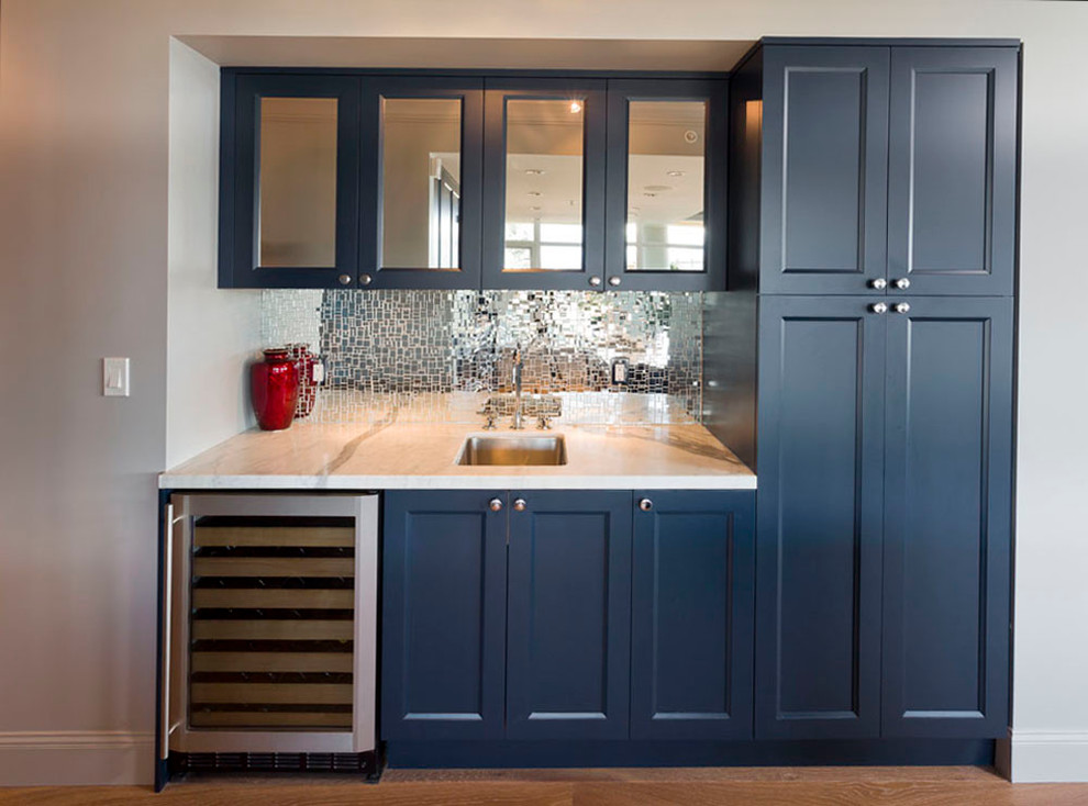 Photo of a mid-sized transitional l-shaped home bar in Vancouver with blue cabinets, medium hardwood floors, an undermount sink, recessed-panel cabinets, marble benchtops, white splashback, marble splashback and brown floor.