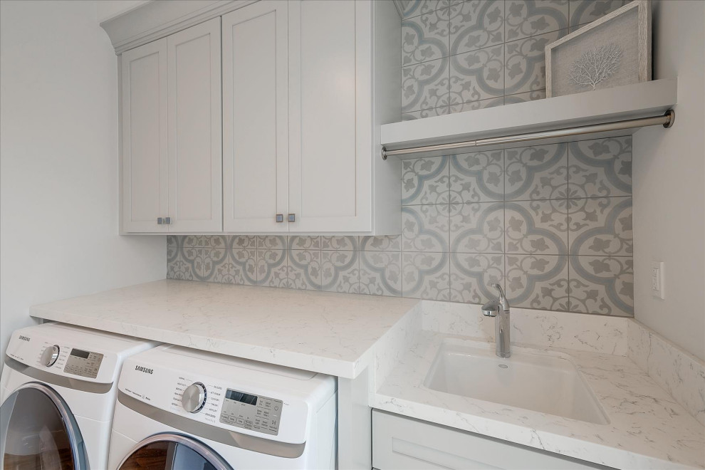 サンフランシスコにある高級な中くらいなビーチスタイルのおしゃれな洗濯室 (ll型、アンダーカウンターシンク、フラットパネル扉のキャビネット、グレーのキャビネット、クオーツストーンカウンター、青いキッチンパネル、セメントタイルのキッチンパネル、白い壁、クッションフロア、左右配置の洗濯機・乾燥機、ベージュの床、グレーのキッチンカウンター) の写真