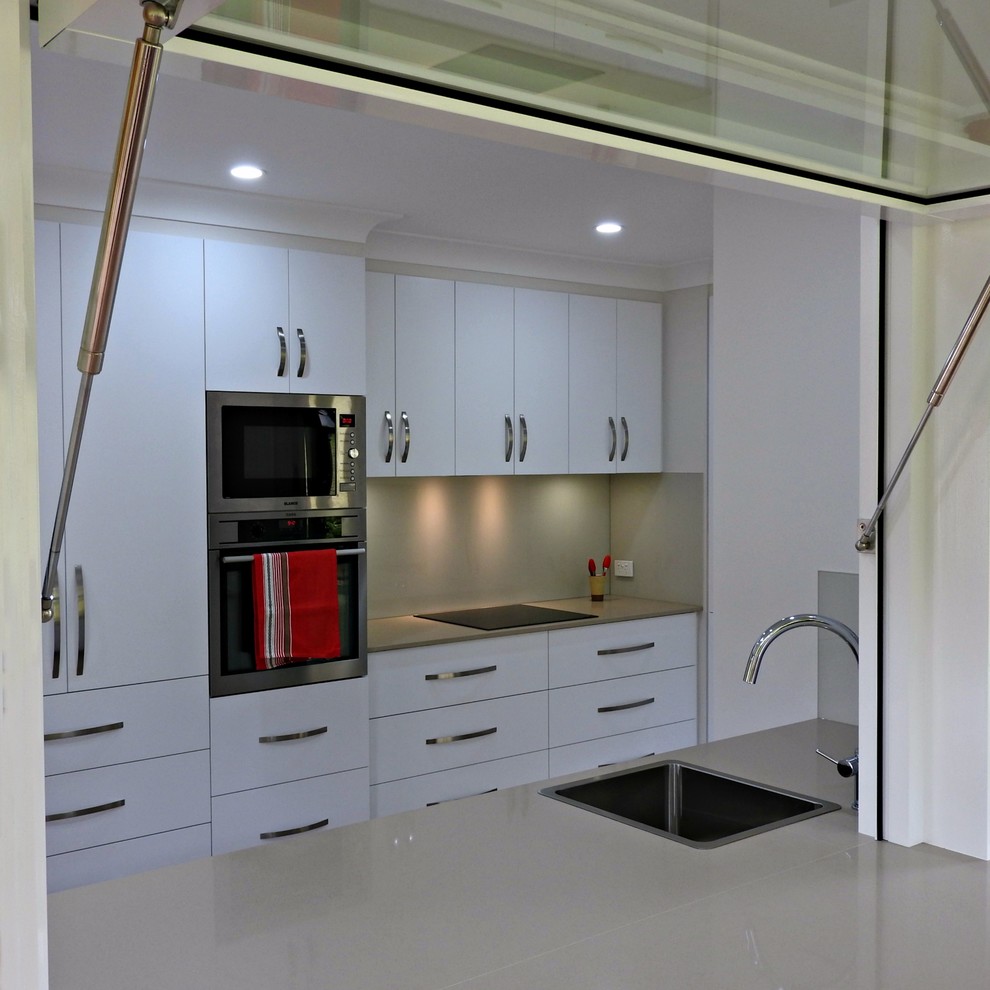 Photo of a mid-sized contemporary galley kitchen pantry in Brisbane with a single-bowl sink, flat-panel cabinets, white cabinets, quartz benchtops, beige splashback, glass sheet splashback, stainless steel appliances, vinyl floors, a peninsula and brown floor.