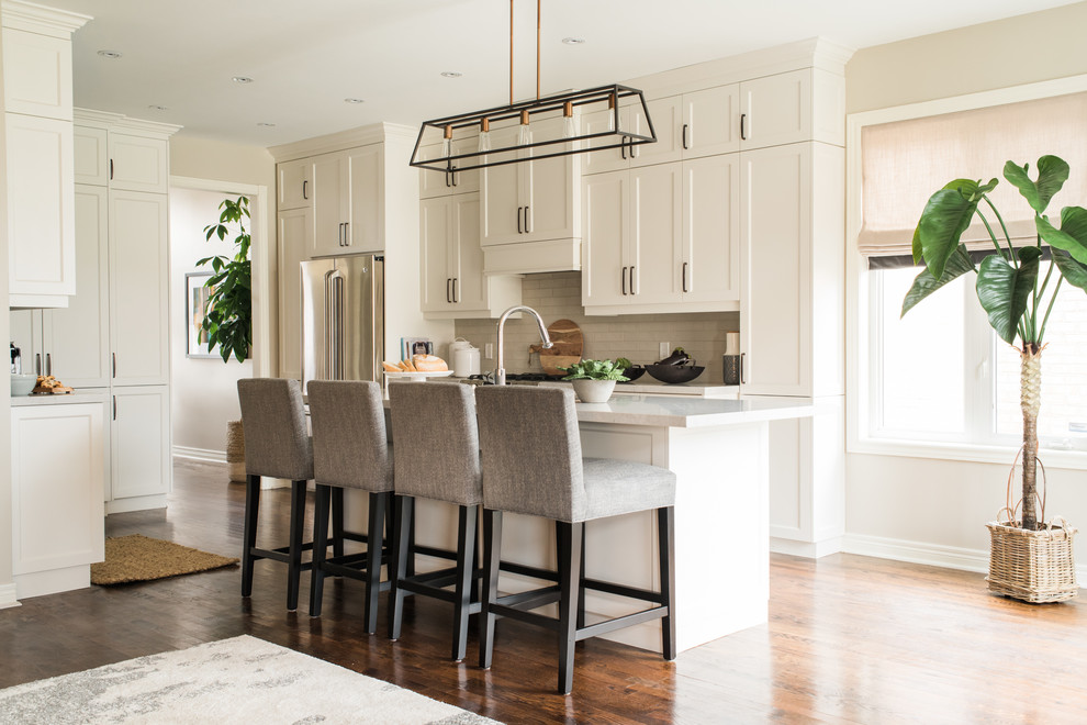 This is an example of a transitional u-shaped open plan kitchen in Toronto with an undermount sink, white cabinets, quartz benchtops, white splashback, porcelain splashback, stainless steel appliances, with island, brown floor, shaker cabinets, dark hardwood floors and white benchtop.